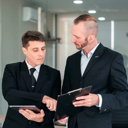 Two men in suits are discussing something, one of them is holding a clipboard. Scene is professional and serious